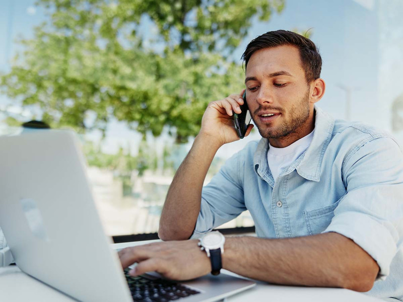 Junger Mann arbeitet draußen an einem Laptop und telefoniert, symbolisiert mobiles Arbeiten und flexible Arbeitsmodelle wie Homeoffice