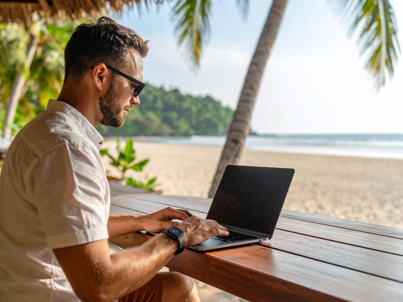 Digitaler Nomade arbeitet im Homeoffice am Strand mit Laptop – flexibles Arbeiten und Work-Life-Blending unter Palmen im Zeitalter von Remote Work.