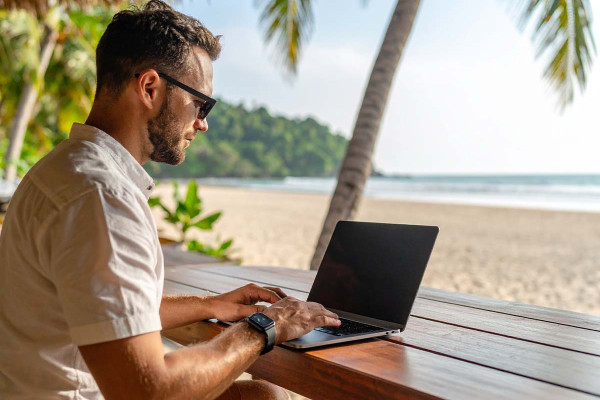 Digitaler Nomade arbeitet im Homeoffice am Strand mit Laptop – flexibles Arbeiten und Work-Life-Blending unter Palmen im Zeitalter von Remote Work.