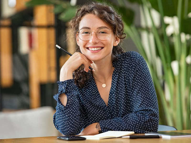 Junge Frau mitten in einer Bibliothek, umgeben von Büchern, vertieft in das Studium der Germanistik. Sie schreibt Notizen in ihr Notizbuch und lächelt dabei, während sie an einem Tisch sitzt.