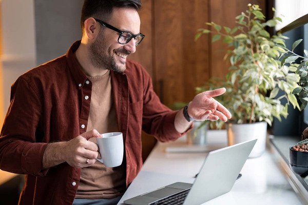 Fröhlicher, selbstbewusster Mann bei hybrider Online-Arbeit oder virtuellem Bewerbungsgespräch im Homeoffice, hält Kaffeetasse und gestikuliert während einer Videokonferenz.