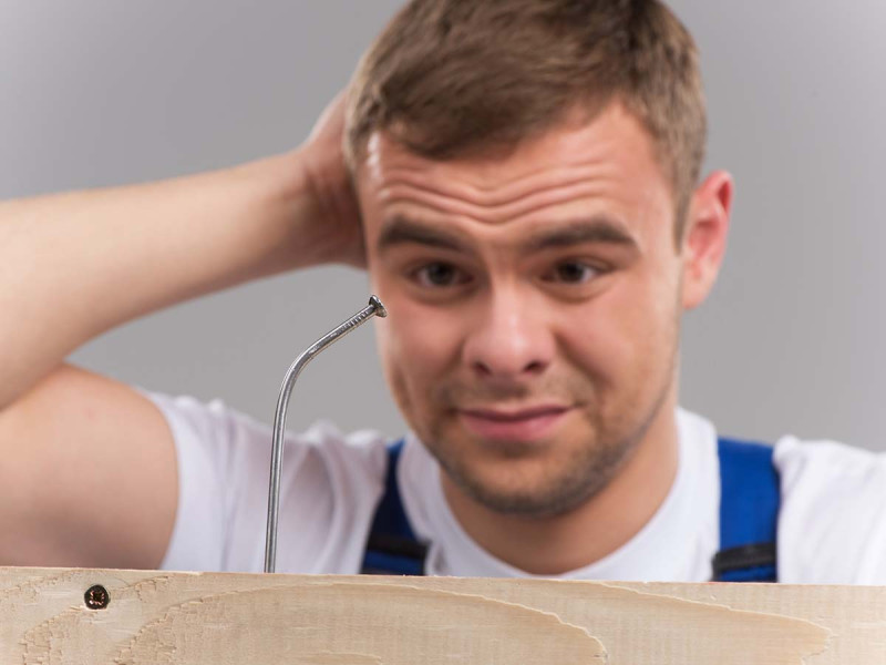 Handwerker betrachtet ratlos einen verbogenen Nagel im Holz, symbolisiert Inkompetenz oder Fehler bei der Arbeit.