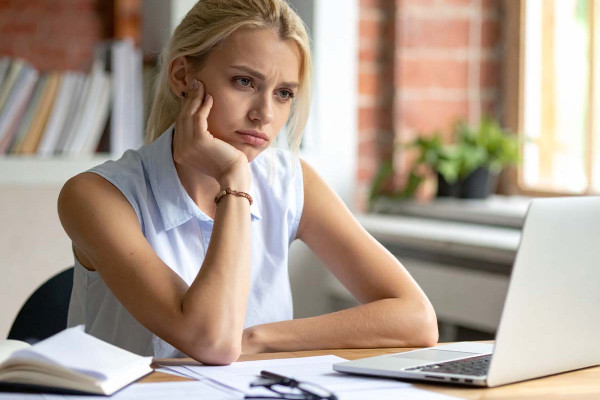 Gestresste junge Frau sitzt am Schreibtisch vor einem Laptop, wirkt erschöpft und nachdenklich, umgeben von Büchern und Arbeitsmaterialien im Homeoffice.