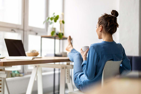 Eine Frau sitzt entspannt mit den Füßen auf dem Tisch und blickt auf ihren ausgeschalteten Computer. Sie wirkt gelassen und hat offensichtlich heute keine Lust auf die Arbeit, von der sie unentschuldigt fernbleibt.