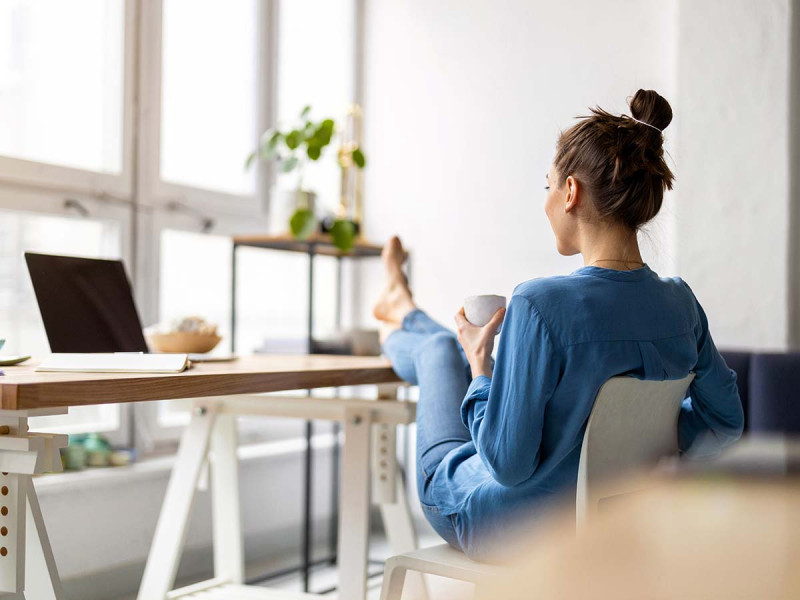 Eine Frau sitzt entspannt mit den Füßen auf dem Tisch und blickt auf ihren ausgeschalteten Computer. Sie wirkt gelassen und hat offensichtlich heute keine Lust auf die Arbeit, von der sie unentschuldigt fernbleibt.