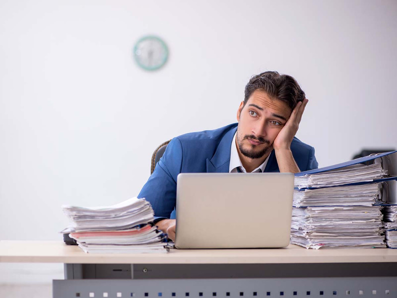 Ungerechte Arbeitsverteilung am Arbeitsplatz: unglücklicher Mitarbeiter sitzt am Schreibtisch und stützt seinen Kopf ab. Er wirft einen fragenden und leeren Blick nach Vorne.