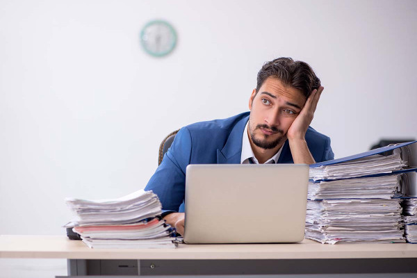 Ungerechte Arbeitsverteilung am Arbeitsplatz: unglücklicher Mitarbeiter sitzt am Schreibtisch und stützt seinen Kopf ab. Er wirft einen fragenden und leeren Blick nach Vorne.