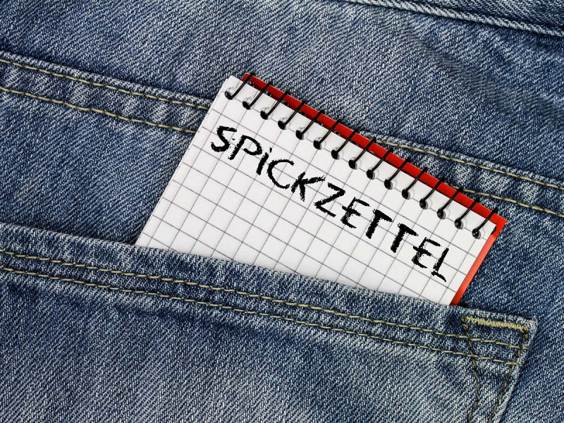 Kariertes Notizbuch mit der Aufschrift ‚Spickzettel‘ ragt aus einer Jeans-Tasche – symbolisch für Spickzettel im Vorstellungsgespräch oder bei Prüfungen