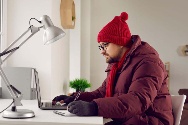Ein Mann arbeitet an einem Schreibtisch mit Laptop und Notizbuch, trägt dabei eine dicke Winterjacke, Mütze, Schal und Handschuhe. Das helle Büro ist schlicht eingerichtet, mit einer Schreibtischlampe und einer kleinen Pflanze im Hintergrund. Das Bild symbolisiert kalte Arbeitsbedingungen und den Versuch, trotz niedriger Temperaturen produktiv zu bleiben.