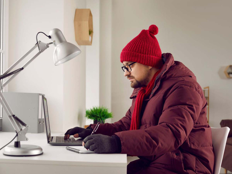 Ein Mann arbeitet an einem Schreibtisch mit Laptop und Notizbuch, trägt dabei eine dicke Winterjacke, Mütze, Schal und Handschuhe. Das helle Büro ist schlicht eingerichtet, mit einer Schreibtischlampe und einer kleinen Pflanze im Hintergrund. Das Bild symbolisiert kalte Arbeitsbedingungen und den Versuch, trotz niedriger Temperaturen produktiv zu bleiben.