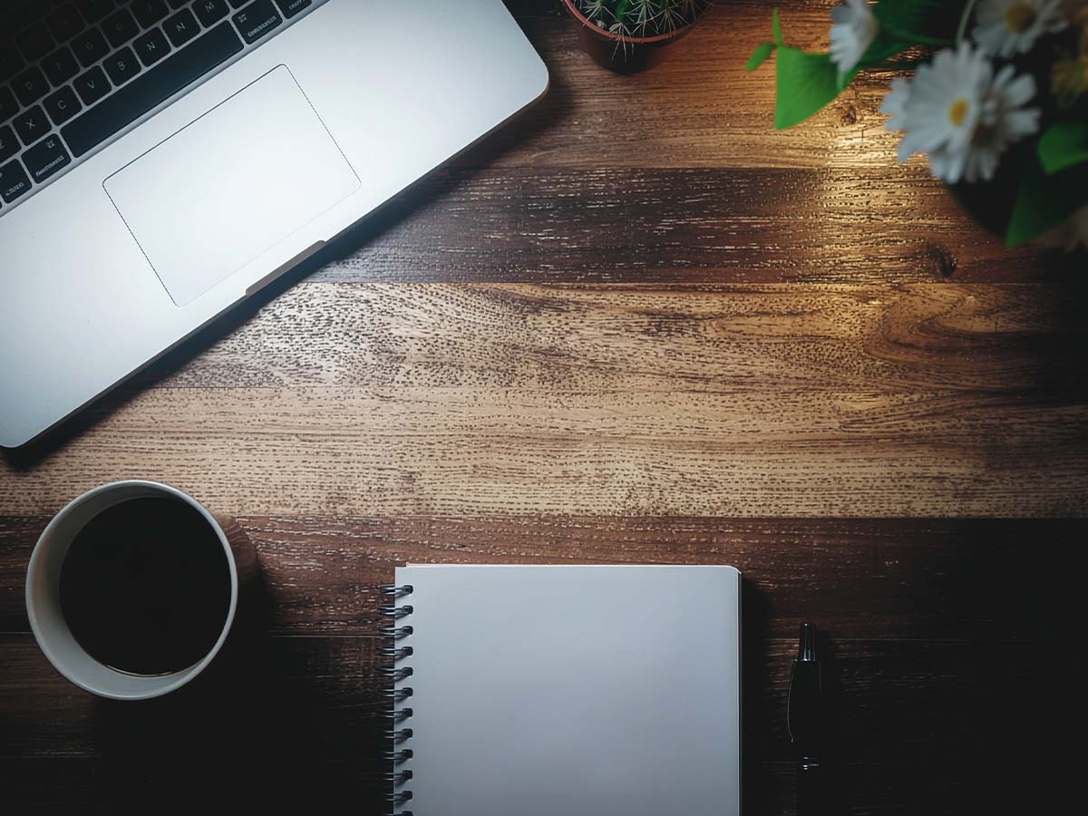 Eine obere Ansicht eines Holzschreibtisches zeigt verschiedene Werkzeuge zur finanziellen Planung, darunter einen einen Laptop mit Investitionsgrafiken, eine Kaffeetasse und ein Notizbuch mit handgeschriebenen Notizen. Die warme Beleuchtung und die organisierte Anordnung der Gegenstände vermitteln eine produktive Arbeitsumgebung.