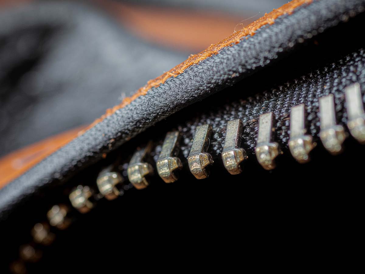 Makroaufnahme eines metallischen Reißverschlusses an einer dunklen Winterjacke mit braunen Stoffnähten. Die Detailaufnahme zeigt die hochwertige Verarbeitung und symbolisiert Schutz, Funktionalität und Strapazierfähigkeit bei winterfester Kleidung.